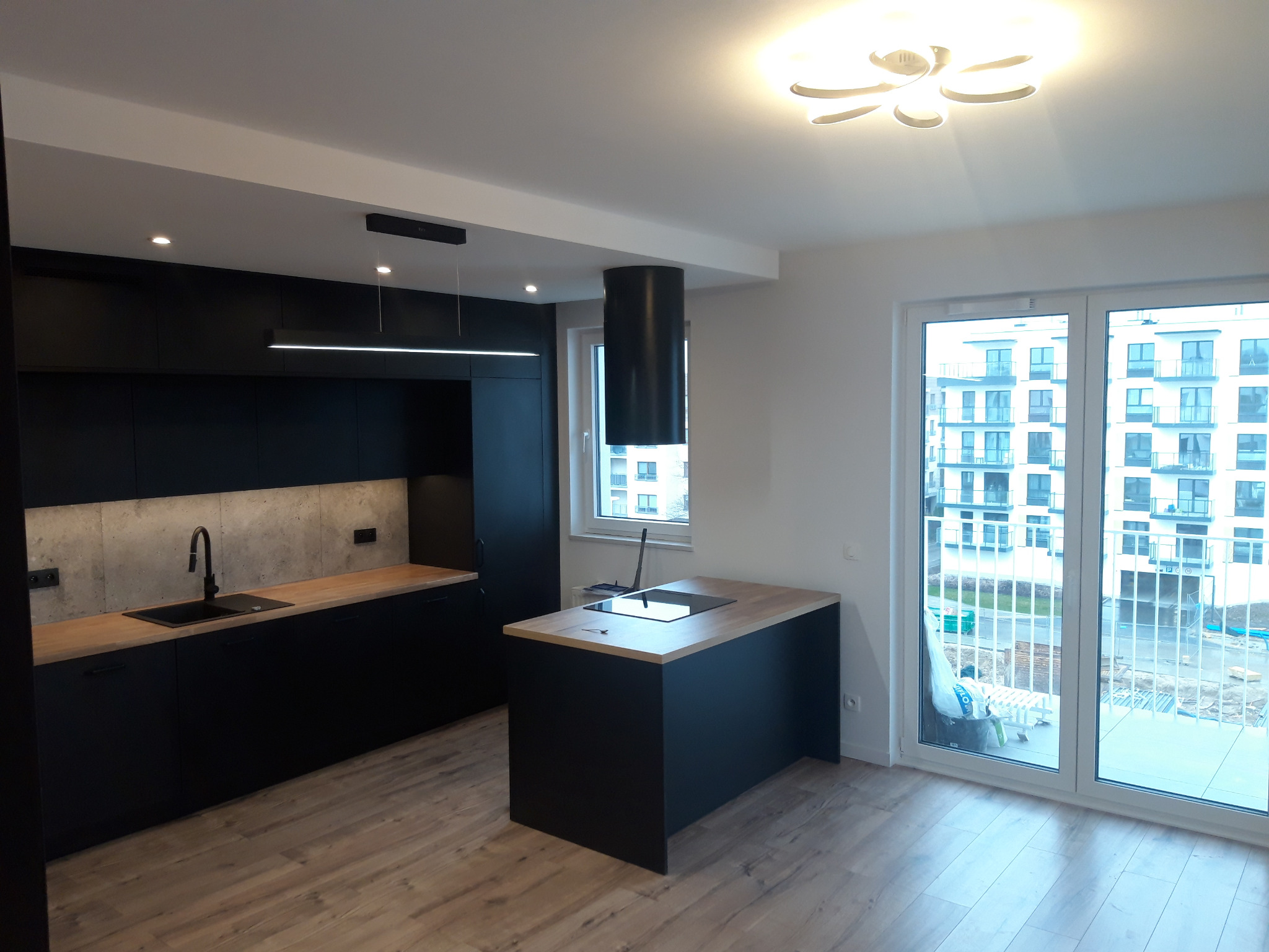 Nowoczesna kuchnia z czarnymi szafkami, drewnianym blatem i wyspą kuchenną z płytą indukcyjną, oświetlona lampą sufitową w kształcie kwiatu i podłużną lampą nad blatem.