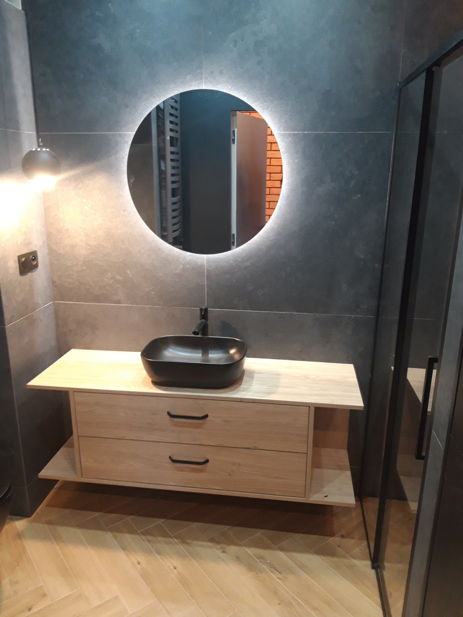 Nowoczesna łazienka z szarymi płytkami, okrągłym lustrem z podświetleniem LED, drewnianą szafką z czarną umywalką nablatową i armaturą w kolorze czarnym, oraz podłogą w jodełkę.