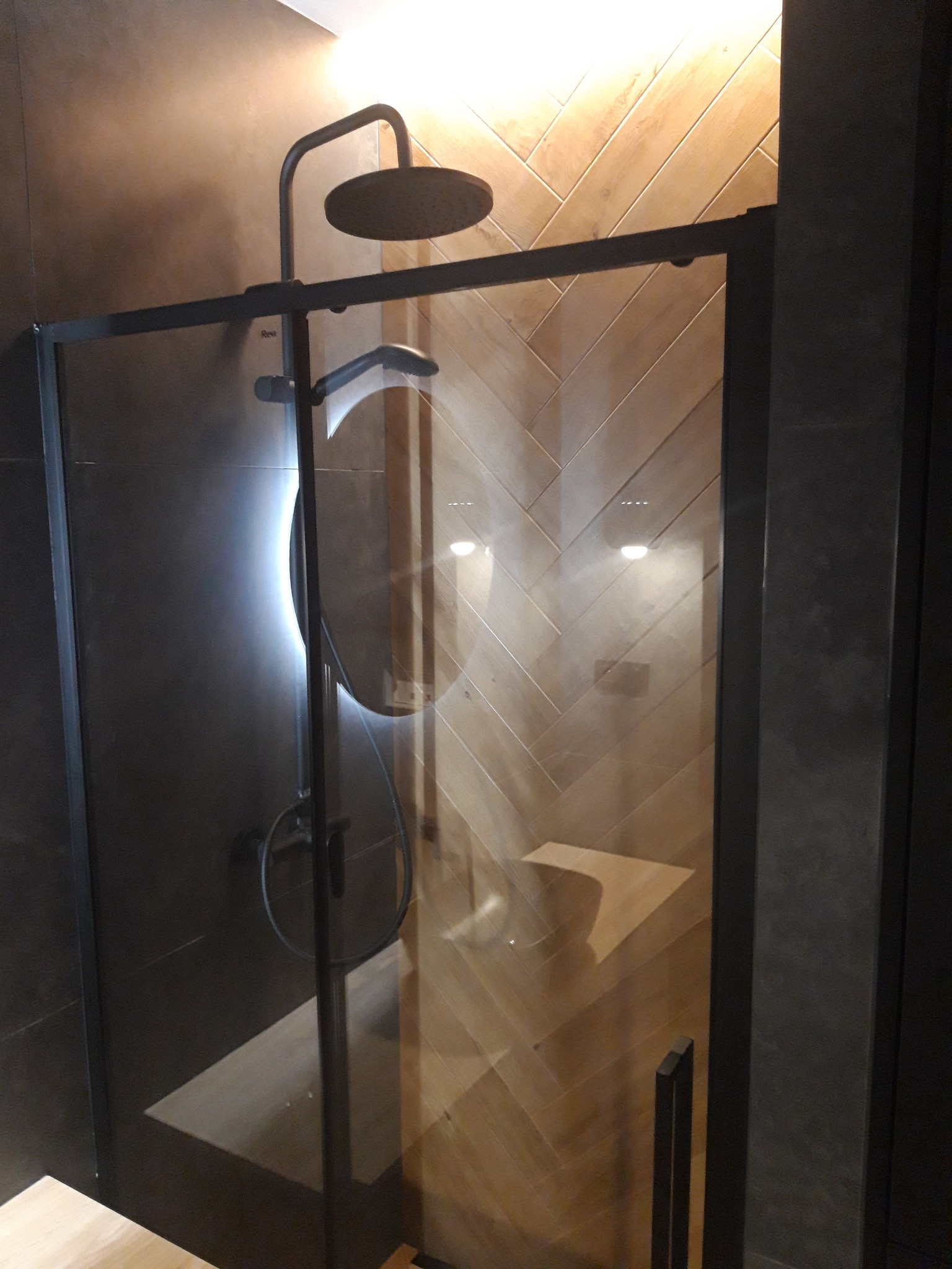 Nowoczesna kabina prysznicowa z czarnymi profilami i deszczownicą, drewnopodobne płytki ułożone w jodełkę na ścianie, widoczne siedzisko wewnątrz kabiny.