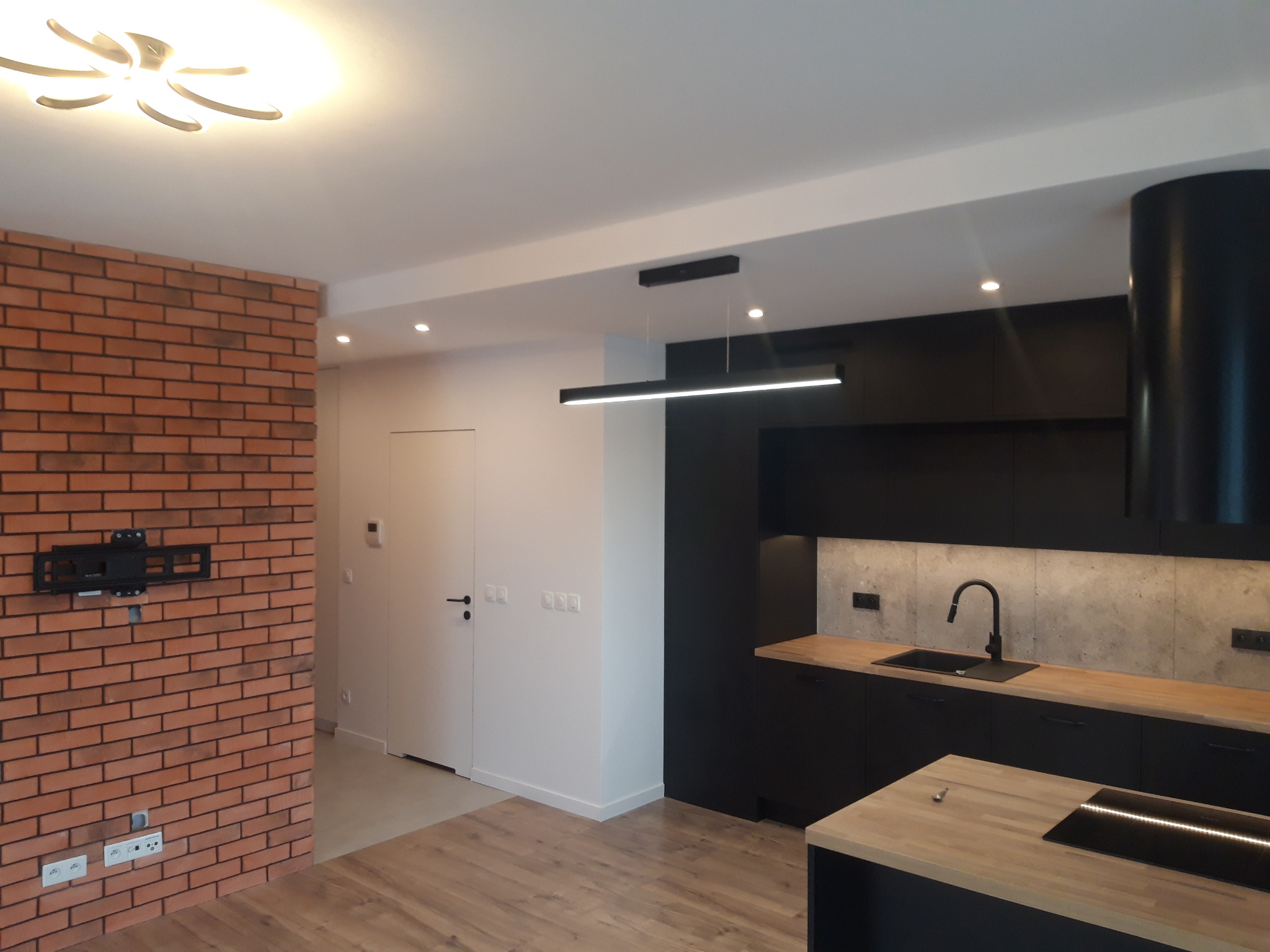 Nowoczesna kuchnia z czarnymi szafkami, drewnianymi blatami i ceglaną ścianą z uchwytem na telewizor, oświetlona designerskimi lampami sufitowymi.