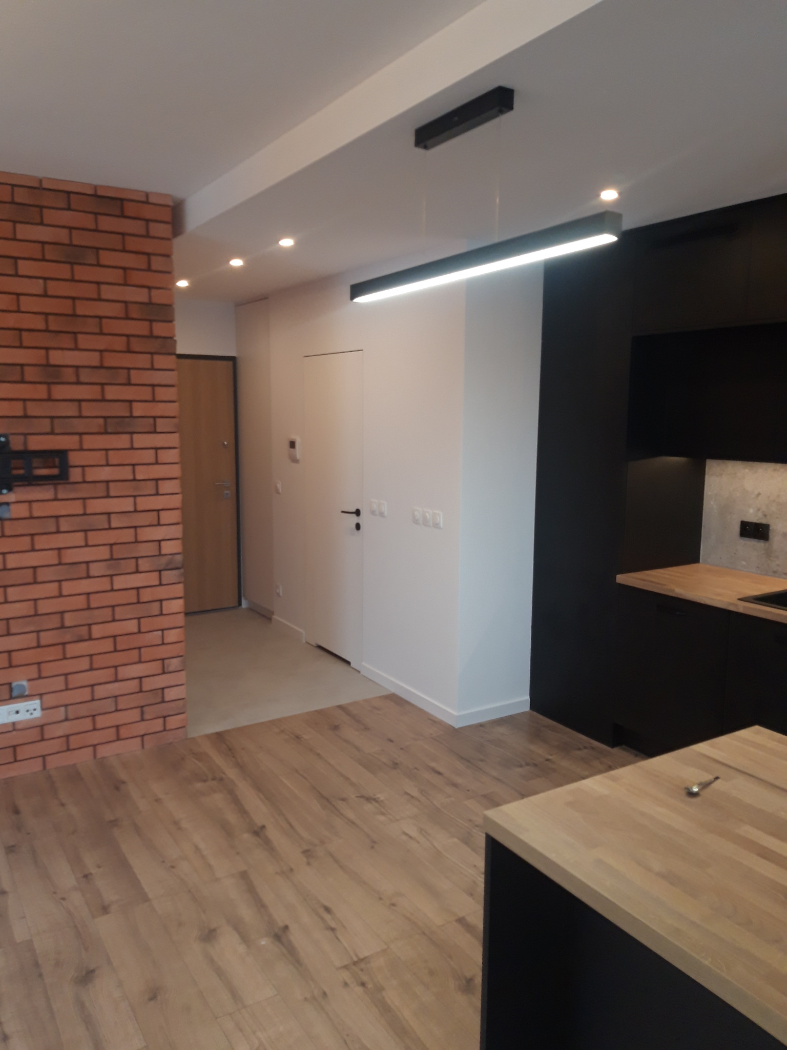Nowoczesne wnętrze mieszkania z ceglaną ścianą, drewnianą podłogą i czarną zabudową kuchenną z drewnianym blatem, oświetlone listwą LED i punktowymi lampami sufitowymi.