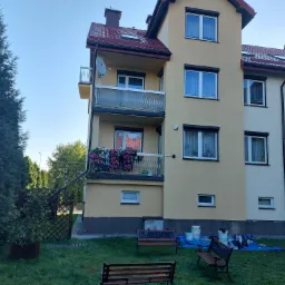 Budynek wielorodzinny po częściowym malowaniu elewacji, widoczne różnice w kolorze, puszki z farbą i zabezpieczenia na trawniku.