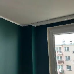 Fragment wnętrza po remoncie: ściana pomalowana na ciemny zielony kolor, biały sufit z listwą ozdobną, okno z roletą w kasecie i widok na budynek mieszkalny.