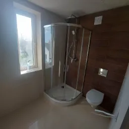 Nowoczesna łazienka z kabiną prysznicową narożną, brązowymi płytkami imitującymi drewno na ścianie, beżowymi płytkami na podłodze i ścianach, oraz wiszącą toaletą.