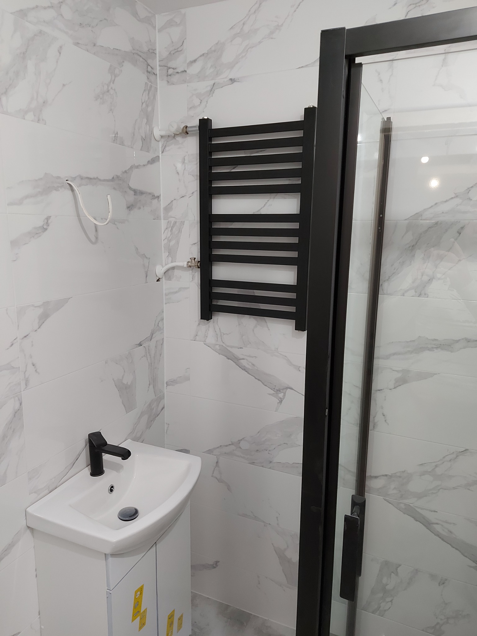 Nowoczesna łazienka z białymi płytkami imitującymi marmur, czarną armaturą i grzejnikiem drabinkowym, kabina prysznicowa z czarnymi profilami.