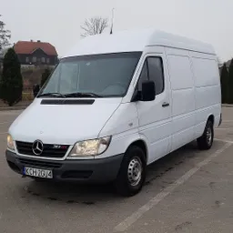 Biały, wysoki furgon Mercedes-Benz Sprinter 313 CDI na parkingu, z widoczną tablicą rejestracyjną KCH 2GU4 i budynkami w tle.