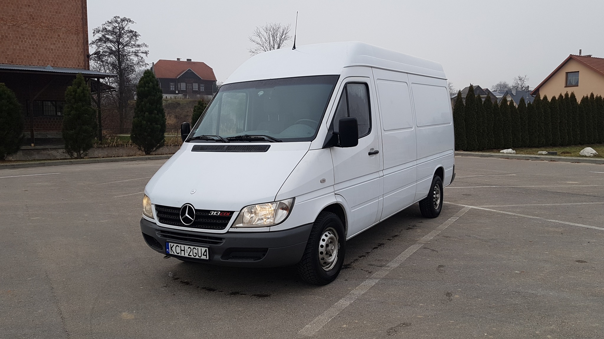 Biały, wysoki furgon Mercedes-Benz Sprinter 313 CDI na parkingu, z widoczną tablicą rejestracyjną KCH 2GU4 i budynkami w tle.