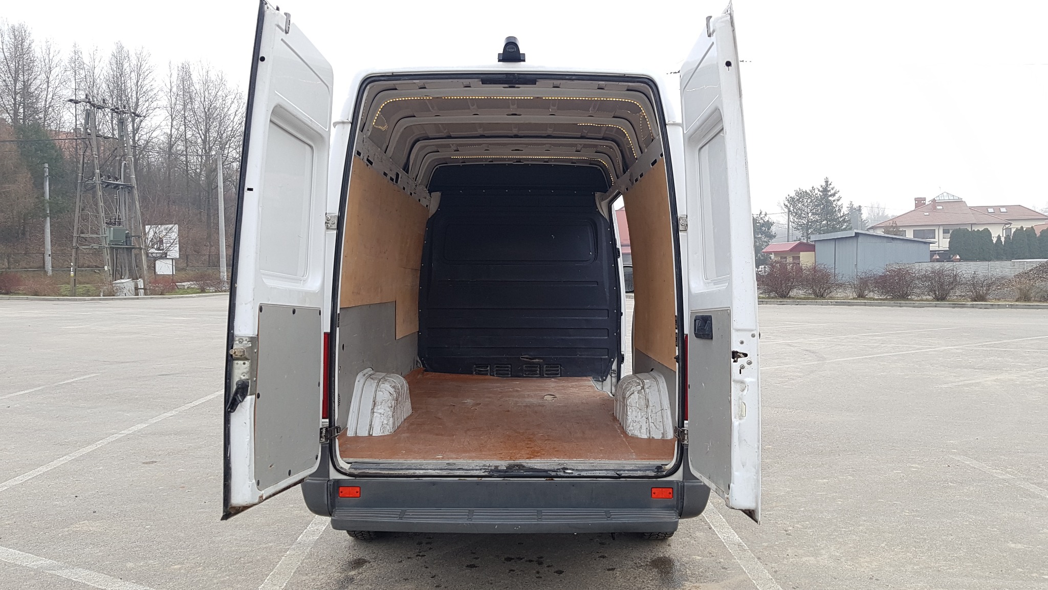 Wnętrze pustego, białego furgonetki z otwartymi tylnymi drzwiami, widok z tyłu na drewnianą podłogę i oświetlenie LED podsufitki.