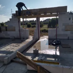 Budowa domu z bloczków betonowych: murarz na drabinie układa nadproże z drewnianych belek, widoczne ściany w trakcie wznoszenia, elementy szalunku i zalana posadzka wewnątrz budynku.