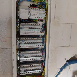 Otwarta skrzynka rozdzielcza Hager z zainstalowanymi bezpiecznikami, różnicówkami i przewodami, zamontowana na surowej ścianie z betonu komórkowego. Widoczny fragment rury karbowanej w ścianie...