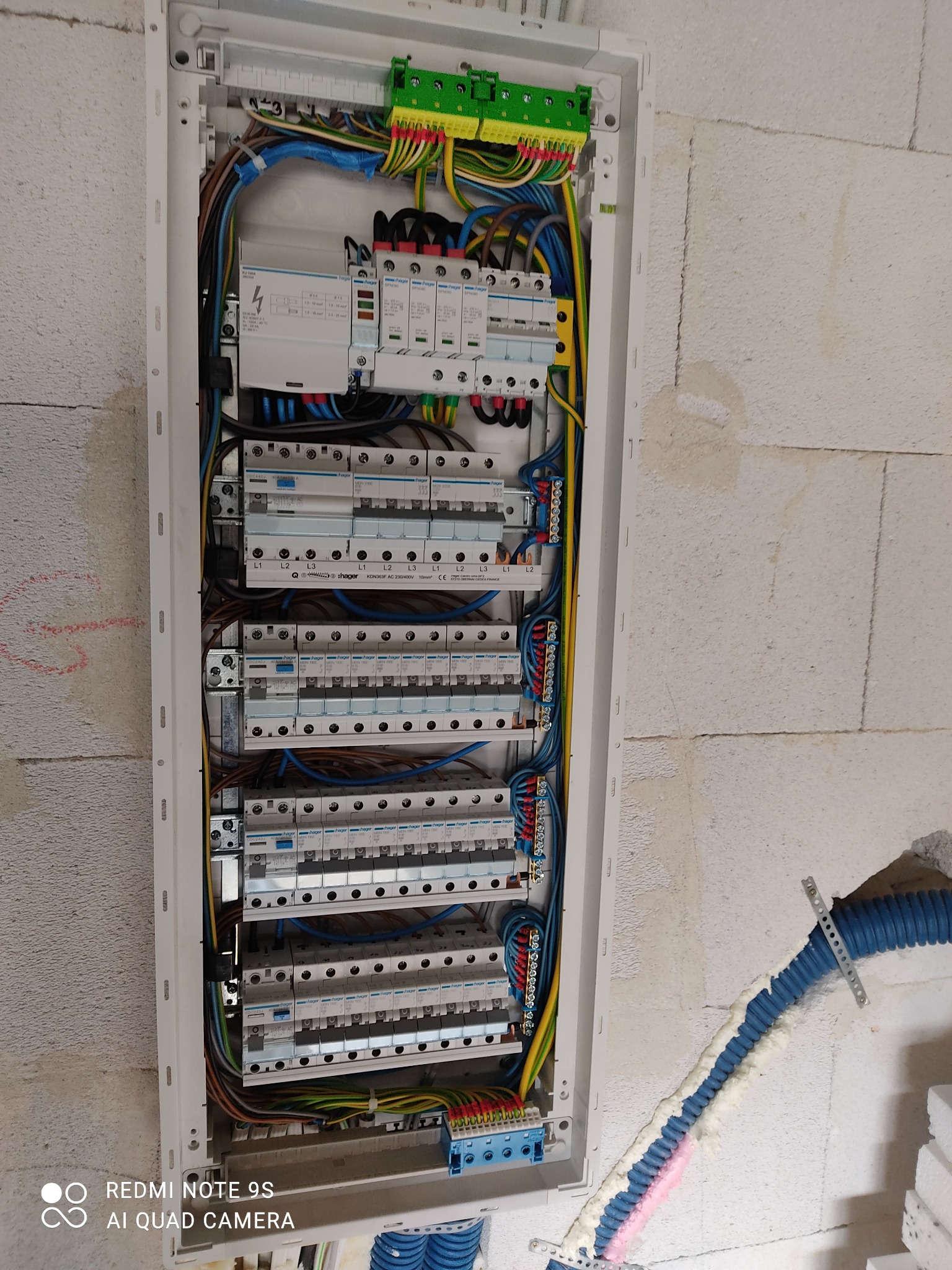 Otwarta skrzynka rozdzielcza Hager z zainstalowanymi bezpiecznikami, różnicówkami i przewodami, zamontowana na surowej ścianie z betonu komórkowego. Widoczny fragment rury karbowanej w ścianie...