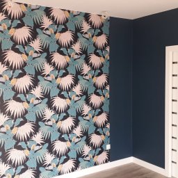 Pro Bau - Pokój po odświeżeniu z granatowymi ścianami, jedna ściana pokryta tapetą w tropikalny wzór z liśćmi palmowymi i kwiatami, białe listwy przypodłogowe i białe drzwi przesuwne z pionowymi wstawkami.