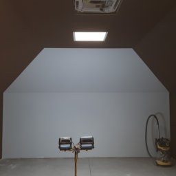 Pro Bau - Poddasze po malowaniu, ściany w dwóch odcieniach brązu i szarości, widoczne narzędzia: lampa oświetleniowa i odkurzacz.