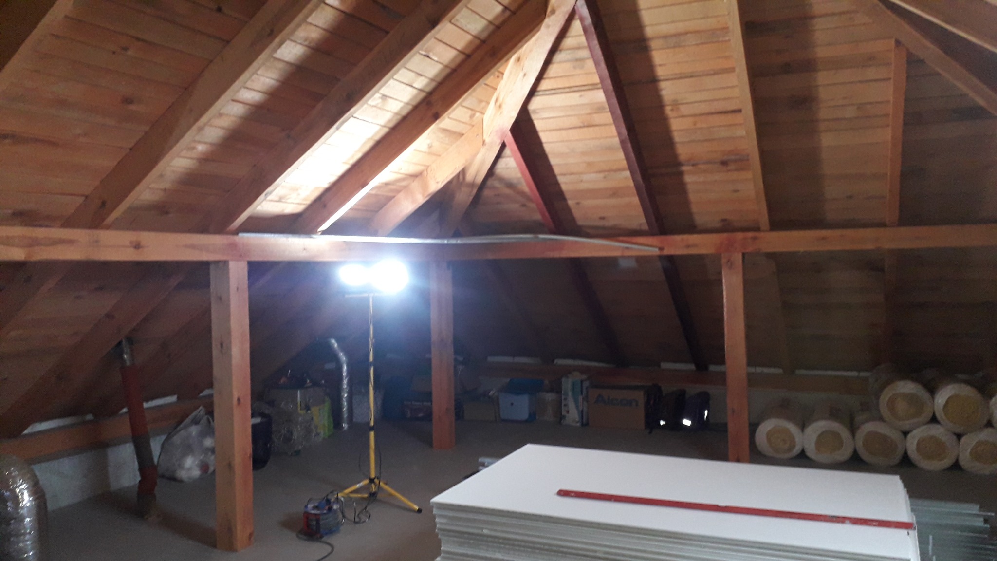 Poddasze z drewnianą konstrukcją dachu, oświetlone lampą budowlaną, z przygotowanymi płytami kartonowo-gipsowymi i materiałami izolacyjnymi, gotowe do montażu.