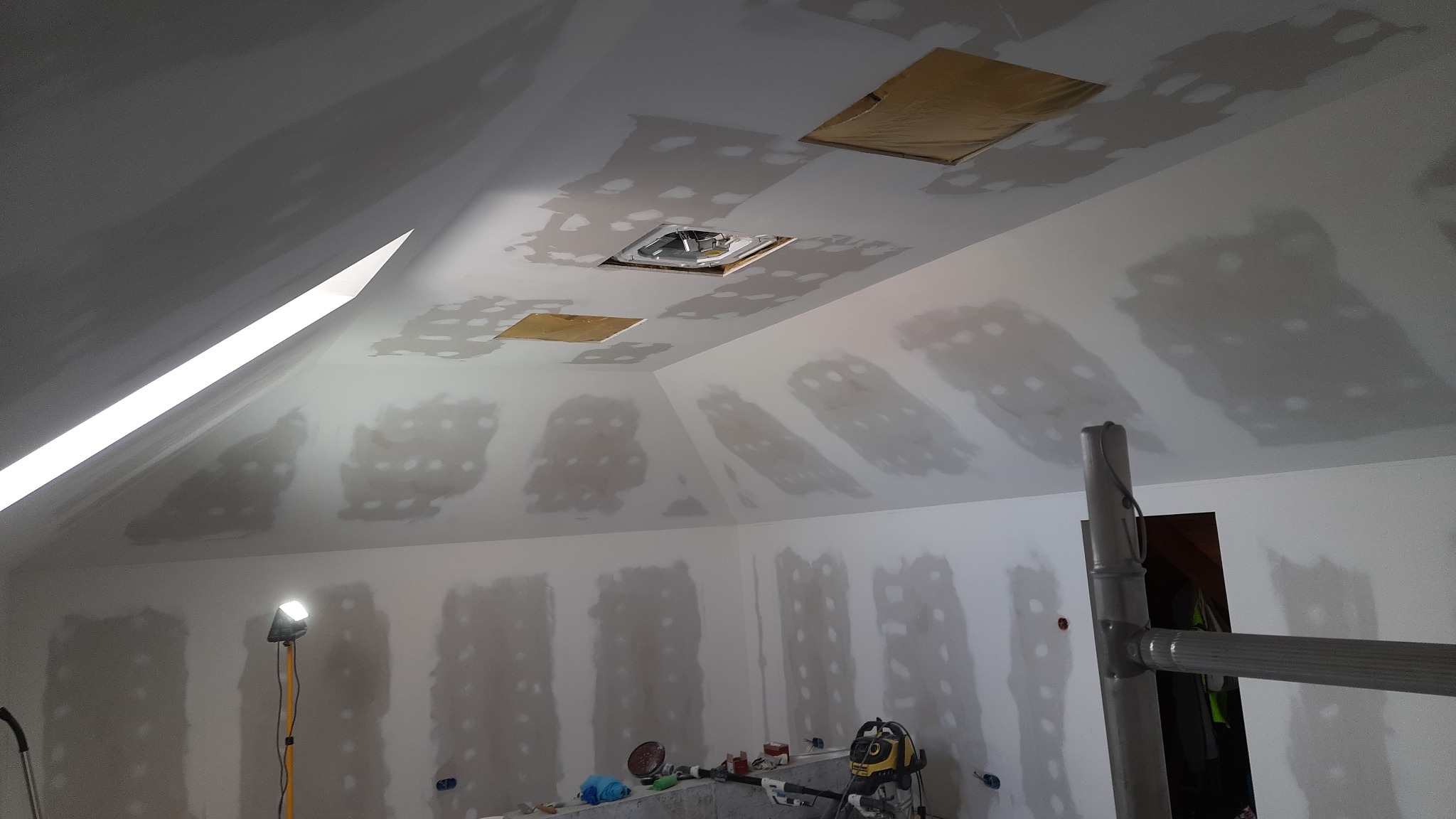 Poddasze w trakcie prac wykończeniowych, widoczne płyty gipsowo-kartonowe na ścianach i suficie ze śladami szpachlowania, otwory na wentylację zabezpieczone folią, lampa robocza na statywie...