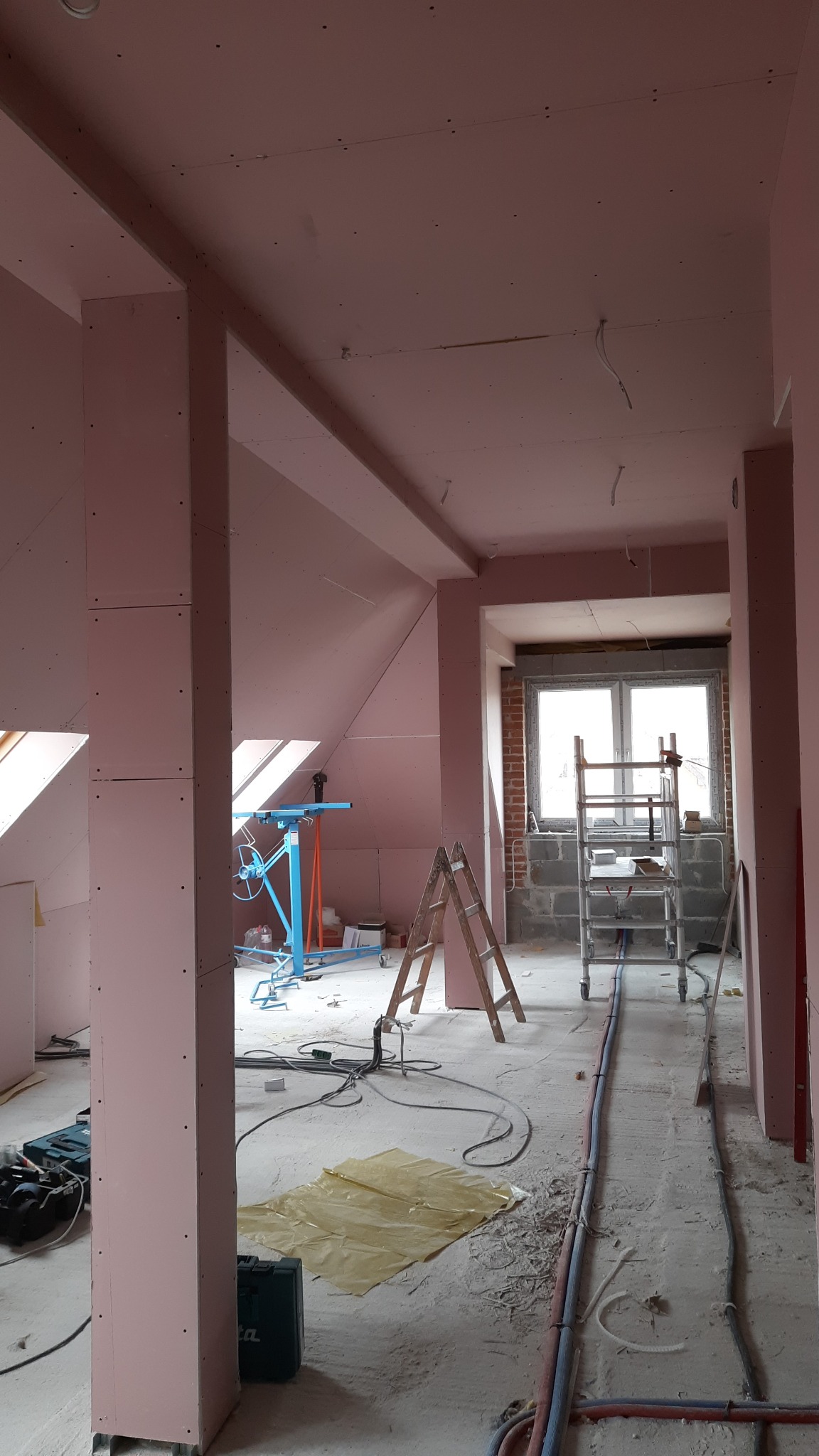 Montaż płyt kartonowo-gipsowych w poddaszu, widoczny podnośnik do płyt, rusztowanie aluminiowe, drewniana drabina, oraz narzędzia na podłodze.