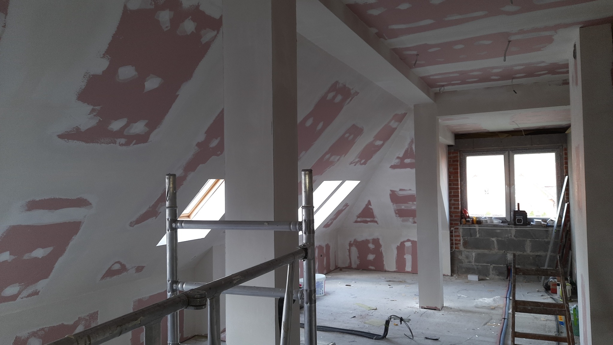 Poddasze w trakcie przygotowania do prac wykończeniowych: widoczne szpachlowane ściany skośne, okna dachowe, rusztowanie oraz okno w murze z widokiem na zewnątrz.