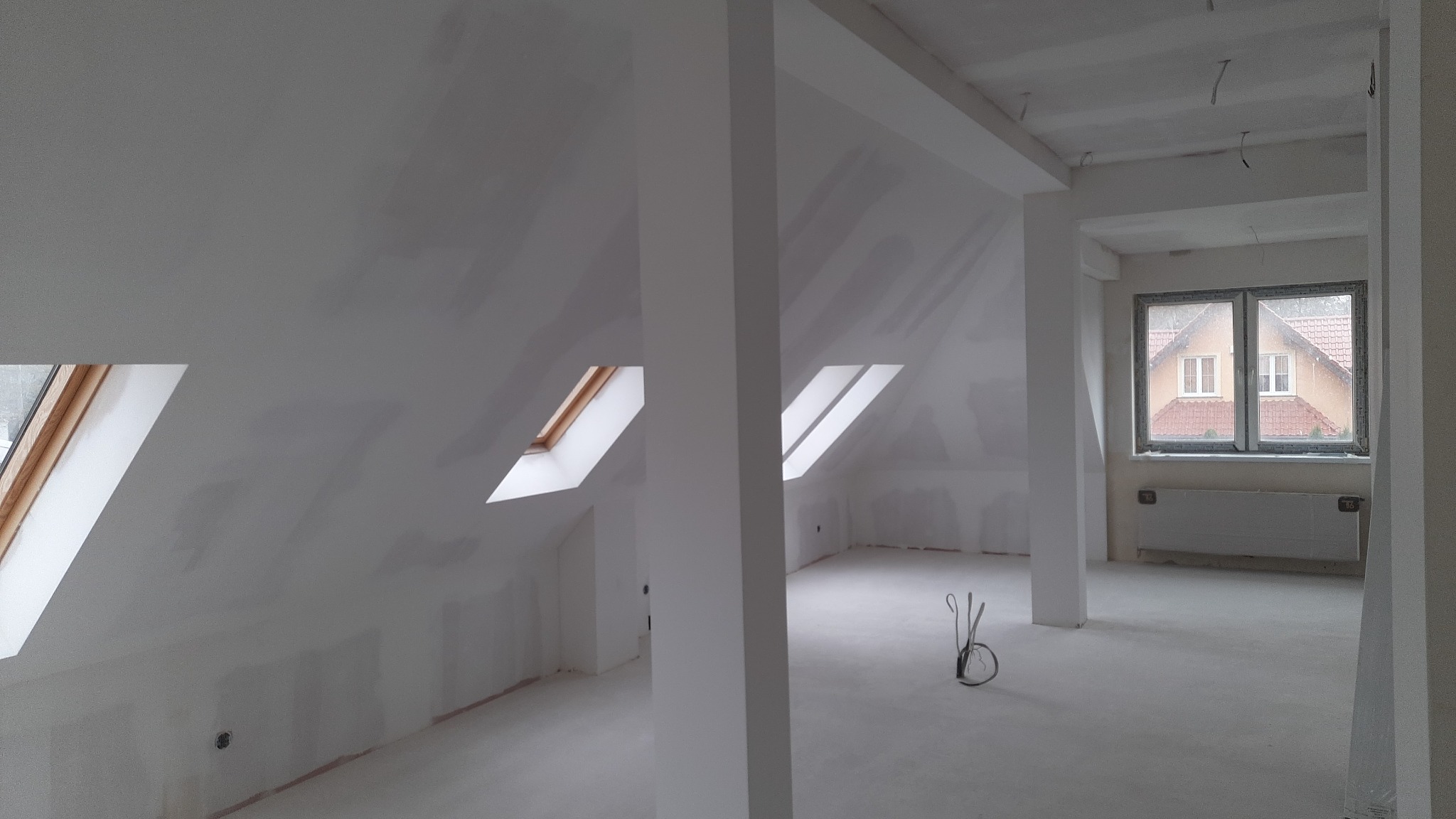 Wnętrze poddasza w trakcie prac wykończeniowych, widoczne okna dachowe, ściany przygotowane do malowania, słupy konstrukcyjne i wystające przewody elektryczne.