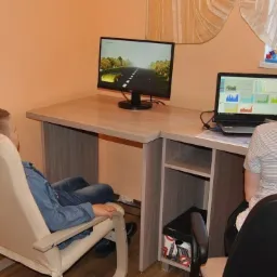 Chłopiec siedzi w fotelu z elektrodami na głowie, obserwując symulację jazdy samochodem na monitorze, podczas gdy kobieta analizuje dane na laptopie.