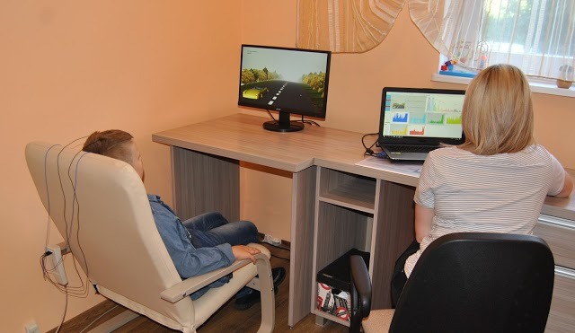 Chłopiec siedzi w fotelu z elektrodami na głowie, obserwując symulację jazdy samochodem na monitorze, podczas gdy kobieta analizuje dane na laptopie.