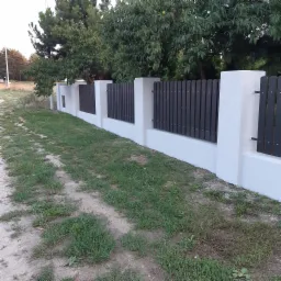 Nowoczesne ogrodzenie z białymi słupkami i ciemnymi, pionowymi deskami, widoczne z perspektywy ścieżki gruntowej.