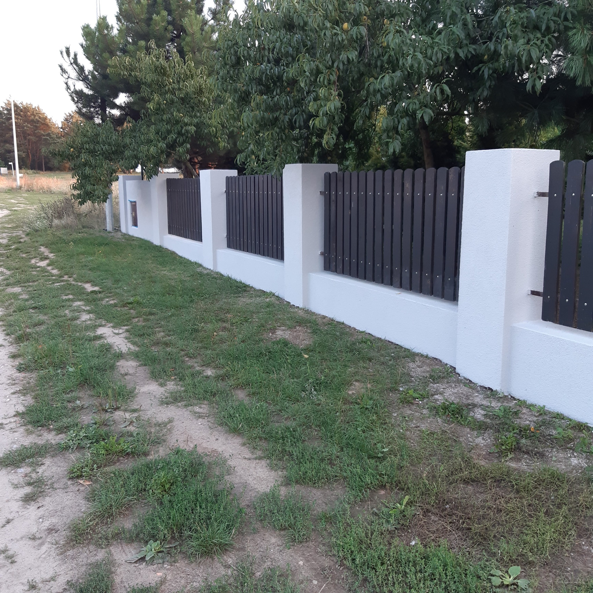 Nowoczesne ogrodzenie z białymi słupkami i ciemnymi, pionowymi deskami, widoczne z perspektywy ścieżki gruntowej.