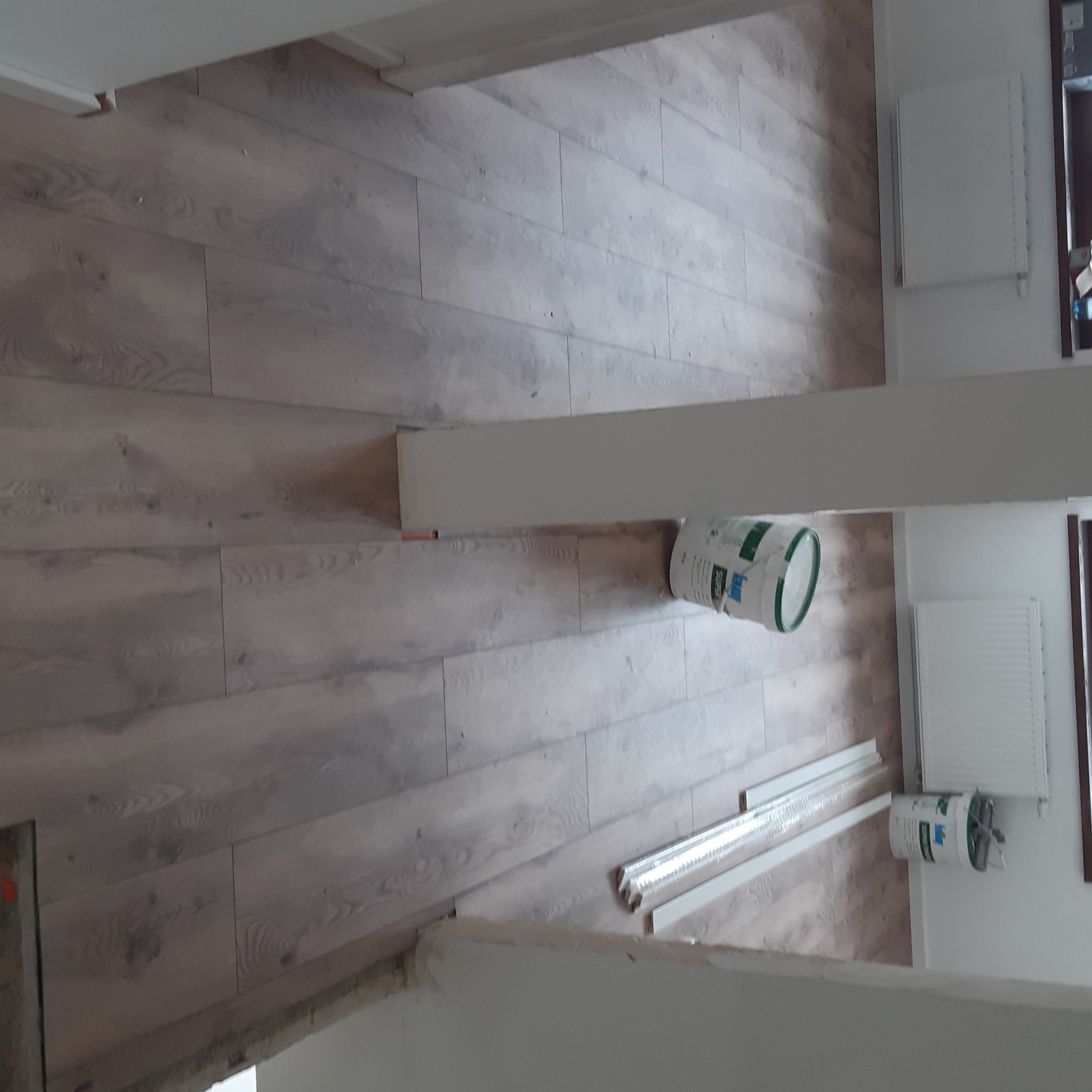 Ułożone panele podłogowe w jasnym odcieniu drewna, widoczny fragment ściany z grzejnikiem, puszka farby i listwy przypodłogowe w folii.