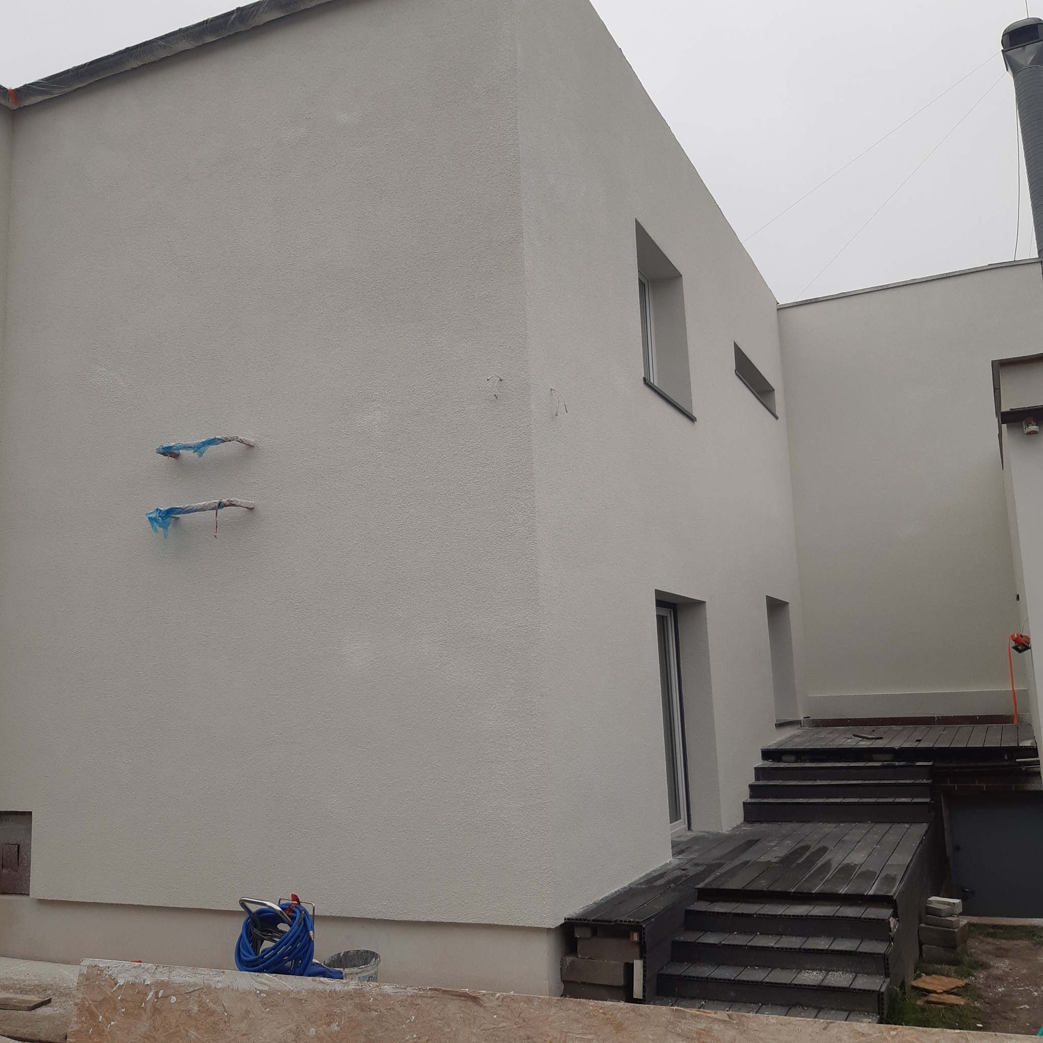 Nowoczesny budynek po wykonaniu elewacji zewnętrznej, widoczne zabezpieczone rurki, osprzęt do malowania i fragment tarasu z kompozytu.