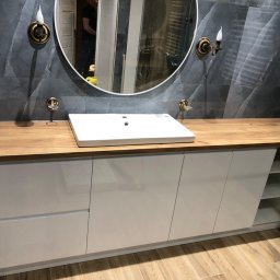 Yomebel-meble na wymiar - Nowoczesna łazienka z drewnianym blatem, białą szafką pod umywalką i okrągłym lustrem z oświetleniem w stylu retro, szare płytki imitujące kamień na ścianie.