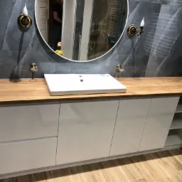 Nowoczesna łazienka z drewnianym blatem, białą szafką pod umywalką i okrągłym lustrem z oświetleniem w stylu retro, szare płytki imitujące kamień na ścianie.