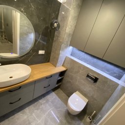 Yomebel-meble na wymiar - Nowoczesna łazienka z szarą marmurową okładziną, drewnianym blatem pod umywalką nablatową, wiszącą szafką i okrągłym lustrem z podświetleniem LED.