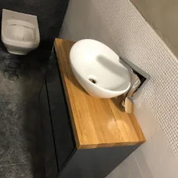 Nowoczesna łazienka z okrągłą, białą umywalką nablatową na blacie z jasnego drewna i czarną szafką, widok z g&oacute;ry, z toaletą w tle.