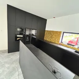 Nowoczesna kuchnia z czarnymi szafkami i wyspą, złote płytki nad blatem, widoczne gniazdko z ładowarką na blacie wyspy, szara podłoga z efektem marmuru.