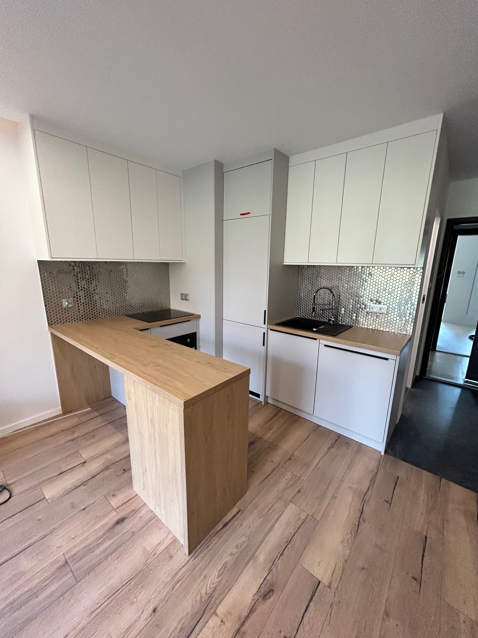 Nowoczesna kuchnia z białymi szafkami i drewnianym blatem, wykończona mozaiką w kształcie plastra miodu na ścianie, z wyspą kuchenną z blatem w tym samym odcieniu drewna.