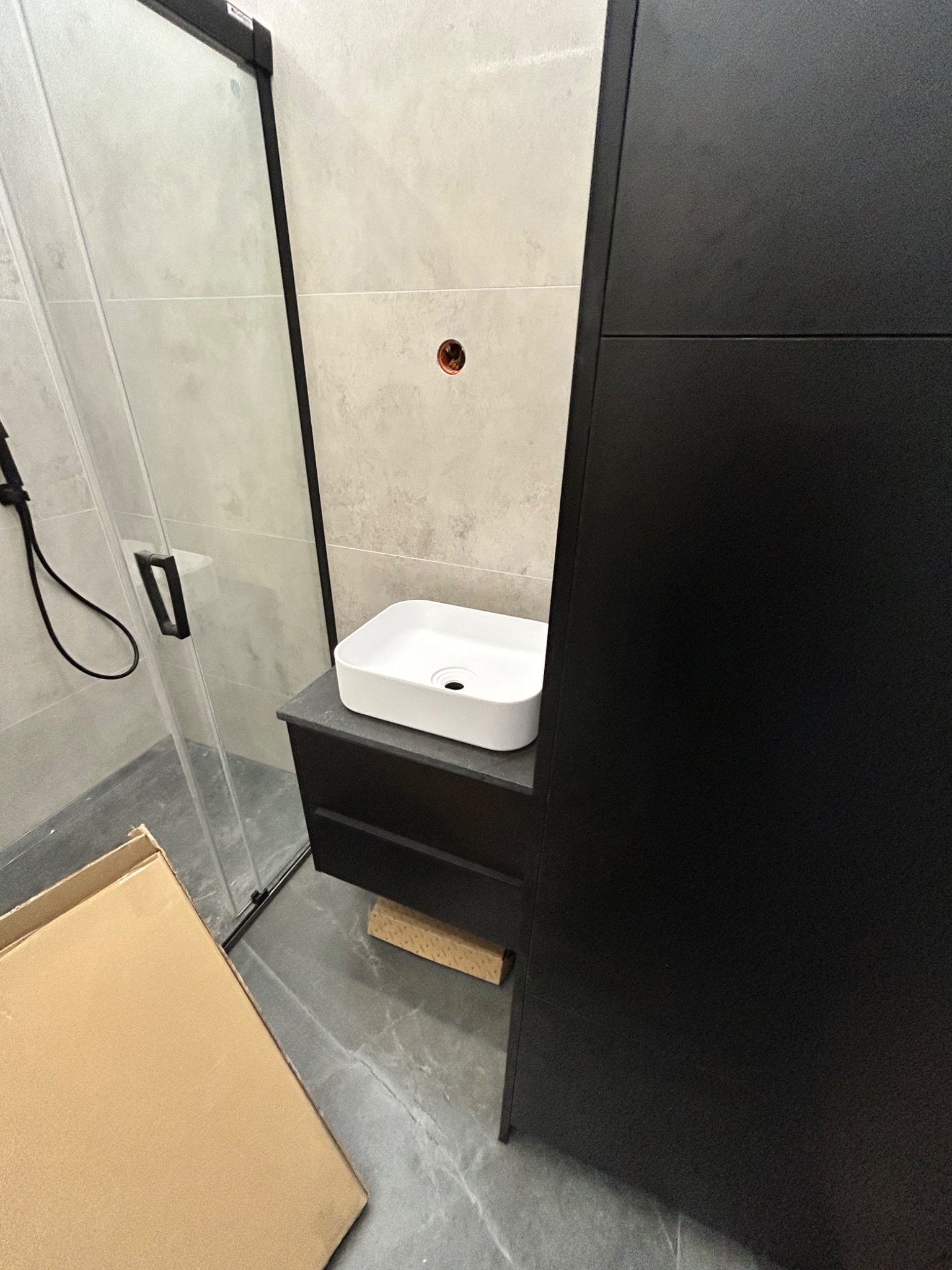 Biała umywalka nablatowa o kwadratowym kształcie zamontowana na czarnej szafce w łazience, widoczna kabina prysznicowa z czarnymi elementami i karton na podłodze.