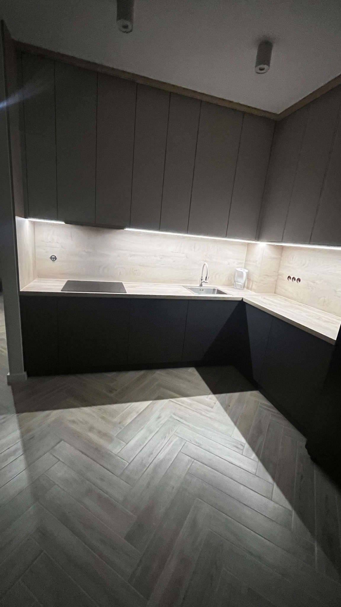 Nowoczesna kuchnia z ciemnymi, gładkimi szafkami, jasnym blatem i oświetleniem LED podszafkowym, uzupełniona podłogą w jodełkę.