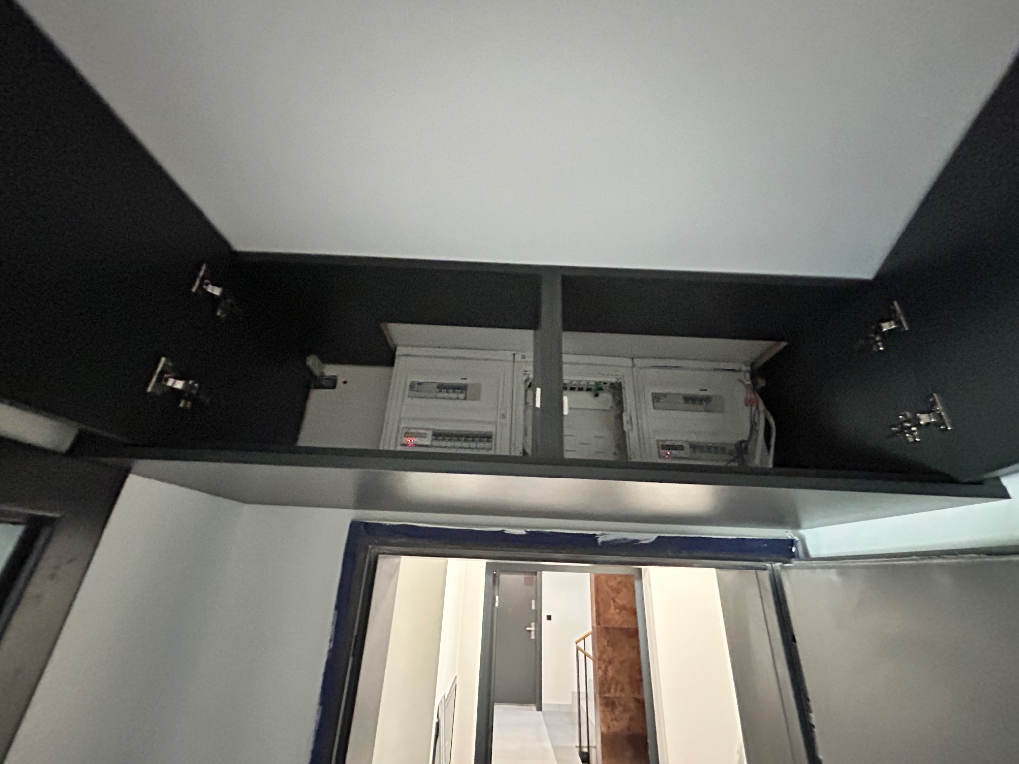 Widok z dołu na szafkę kuchenną z czarnymi frontami, w której ukryte są skrzynki elektryczne, widoczny fragment korytarza w tle.