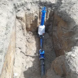 Głęboki wykop w ziemi z zainstalowaną, pionową, niebiesko-czarną rurą wodociągową, częściowo owiniętą białą folią ochronną, widoczne ślady świeżych prac ziemnych i warstwy gleby.