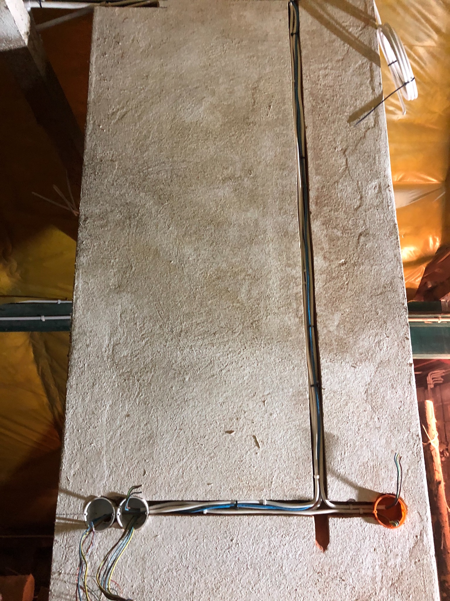 Szara ściana w trakcie prac elektrycznych z widocznymi przewodami w korytkach i puszkach instalacyjnych, izolacja z folii aluminiowej w tle.