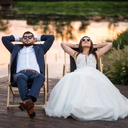 FOTO-KADR - Para młoda w okularach leży na leżakach nad wodą. Pan młody w garniturze, panna młoda w sukni ślubnej. Romantyczna sesja plenerowa w ciepłym świetle zachodzącego słońca.