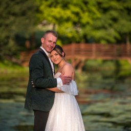 FOTO-KADR - Uśmiechnięta para młoda w objęciach na tle zieleni i drewnianego mostku. Pan młody w zielonym smokingu, panna młoda w białej sukni. Romantyczna sceneria.