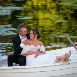 FOTO-KADR - Zakochana para młoda w łódce na jeziorze, on w zielonym smokingu, ona w białej sukni, romantyczna sesja plenerowa, naturalne światło, w tle zielona woda.