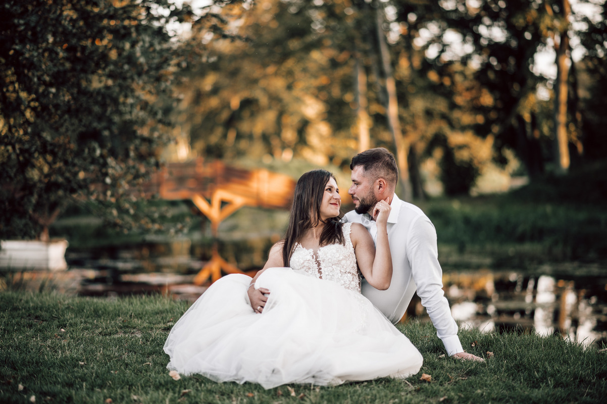 Zakochana para w strojach ślubnych siedzi na trawie nad brzegiem stawu z drewnianym mostkiem w tle, spoglądając na siebie z czułością.