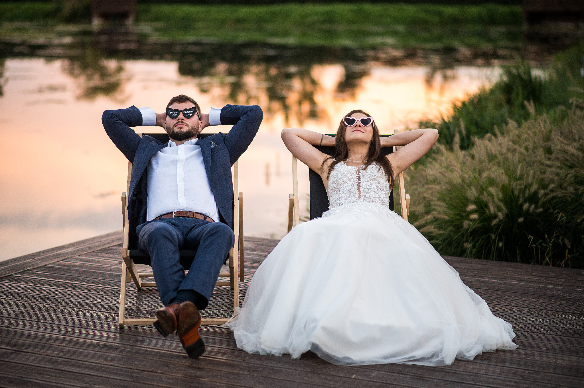 Para młoda w okularach leży na leżakach nad wodą. Pan młody w garniturze, panna młoda w sukni ślubnej. Romantyczna sesja plenerowa w ciepłym świetle zachodzącego słońca.