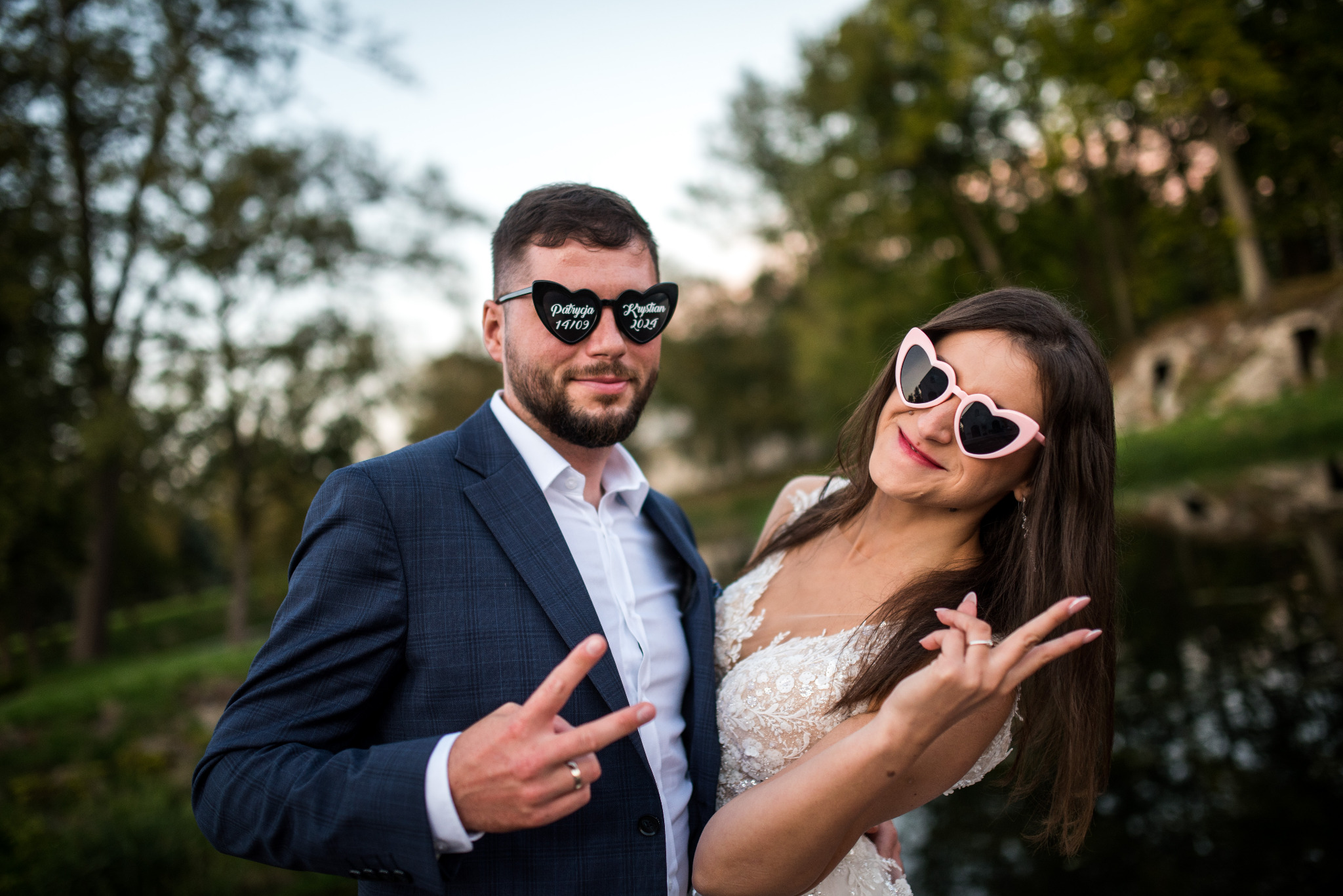 Para młoda w okularach w kształcie serc z datami ślubu, pozuje na tle zieleni. Pan młody w garniturze, panna młoda w koronkowej sukni.