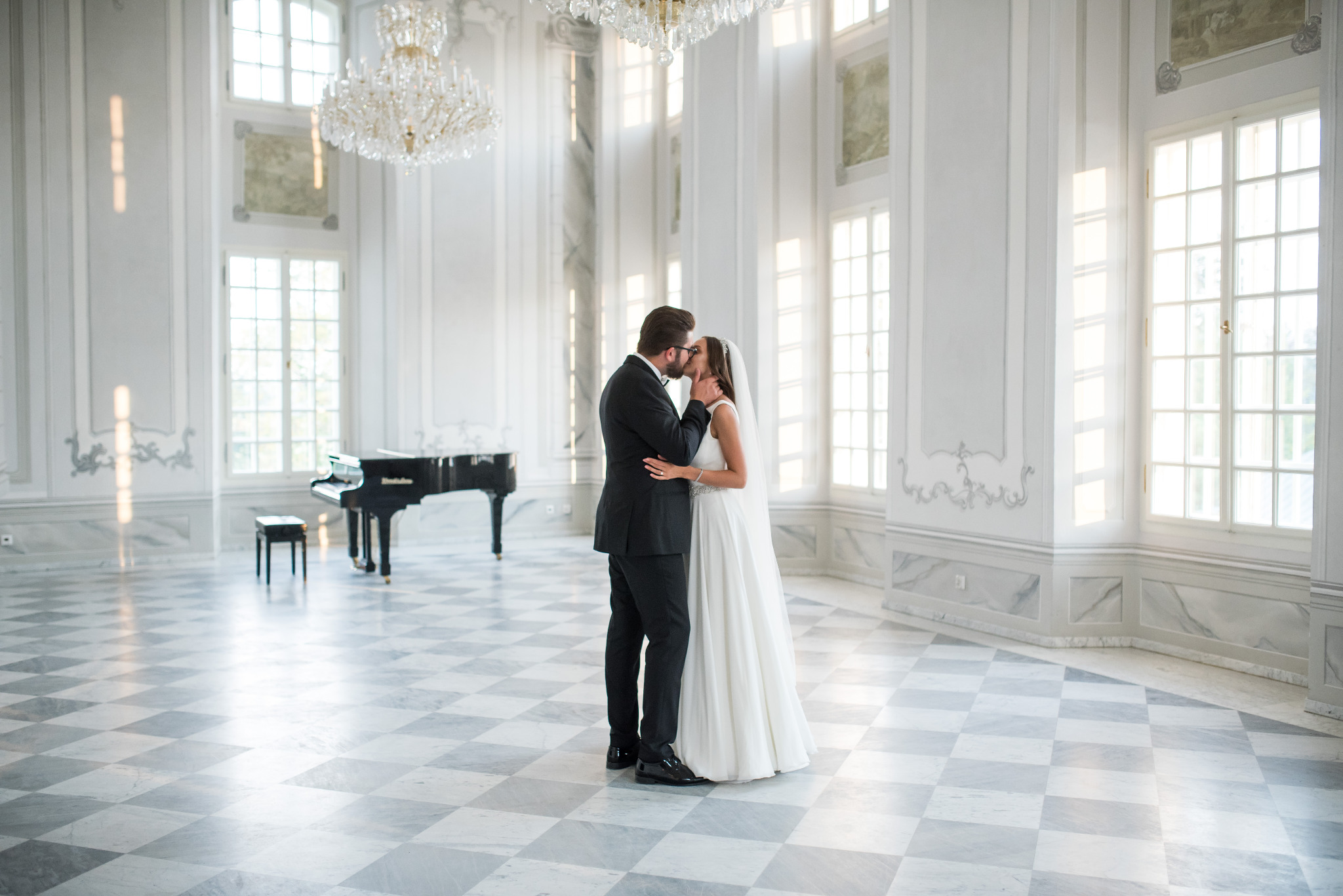 Para młoda w eleganckim wnętrzu, pocałunek na tle fortepianu i kryształowych żyrandoli. Marmurowa podłoga w szachownicę, wysokie okna z delikatnym światłem.