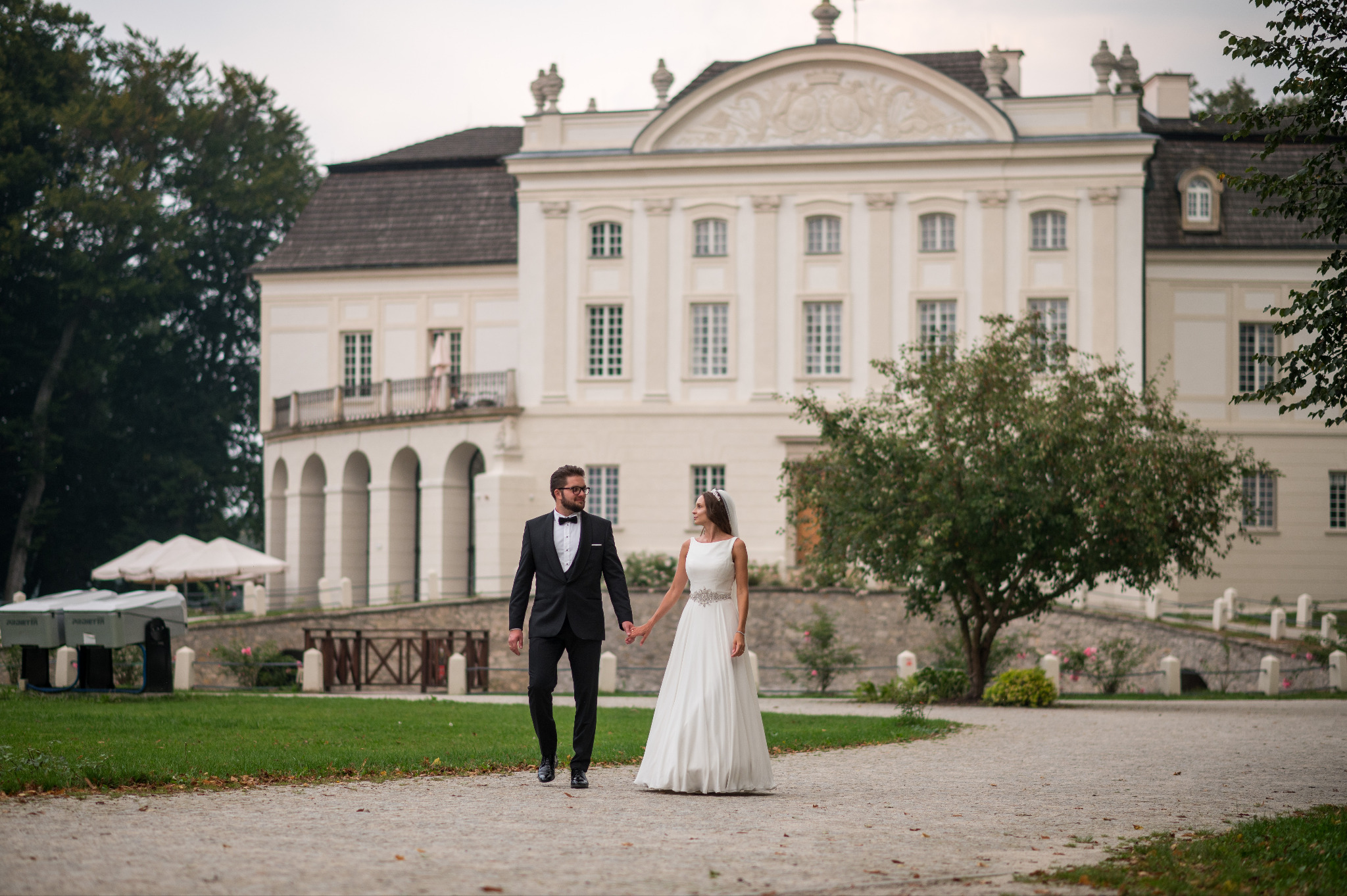 Para młoda spaceruje po parku na tle pałacu. Pan młody w czarnym smokingu, panna młoda w białej sukni. Romantyczna sceneria, delikatne światło, piękna architektura.