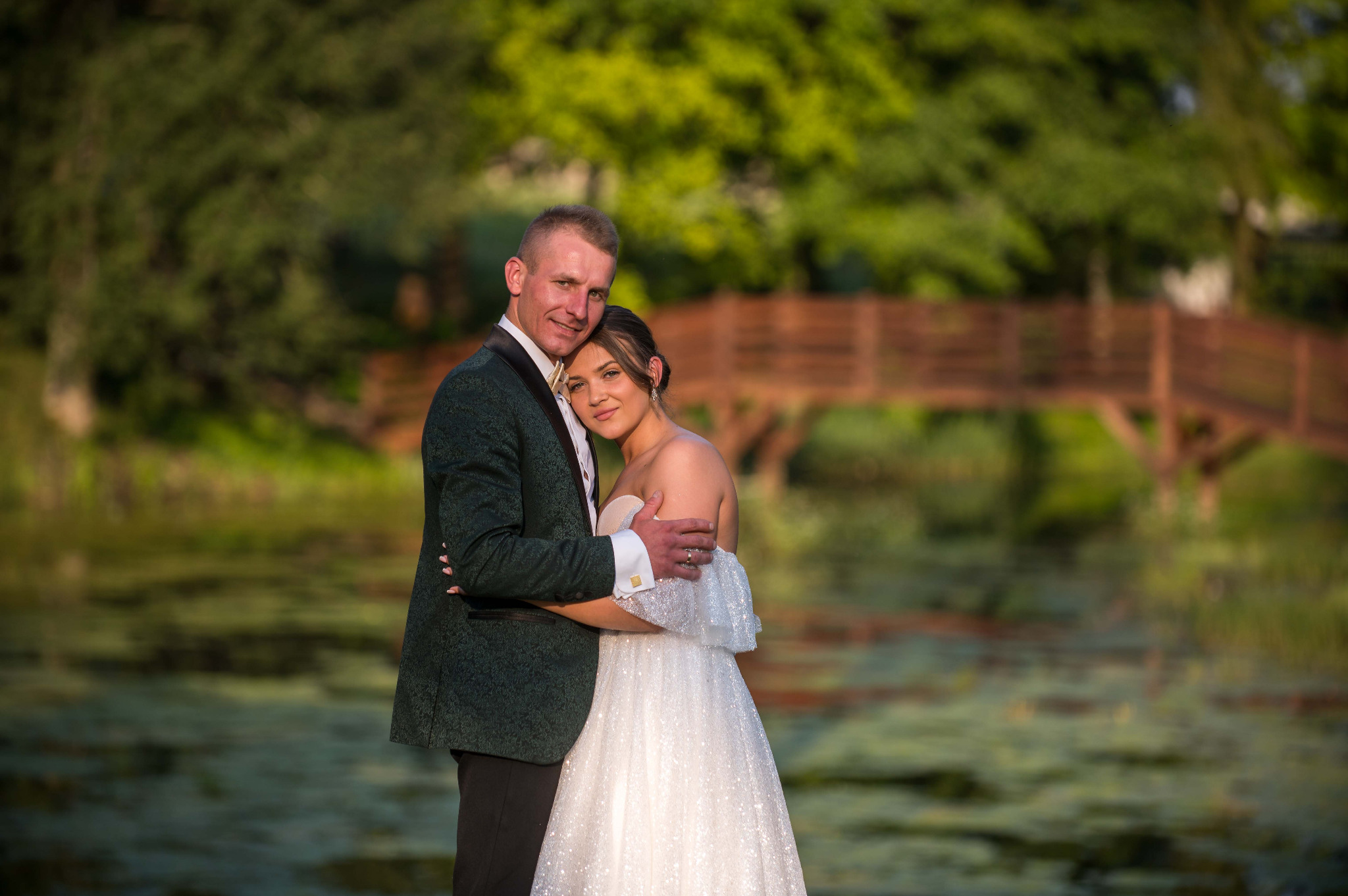 Uśmiechnięta para młoda w objęciach na tle zieleni i drewnianego mostku. Pan młody w zielonym smokingu, panna młoda w białej sukni. Romantyczna sceneria.