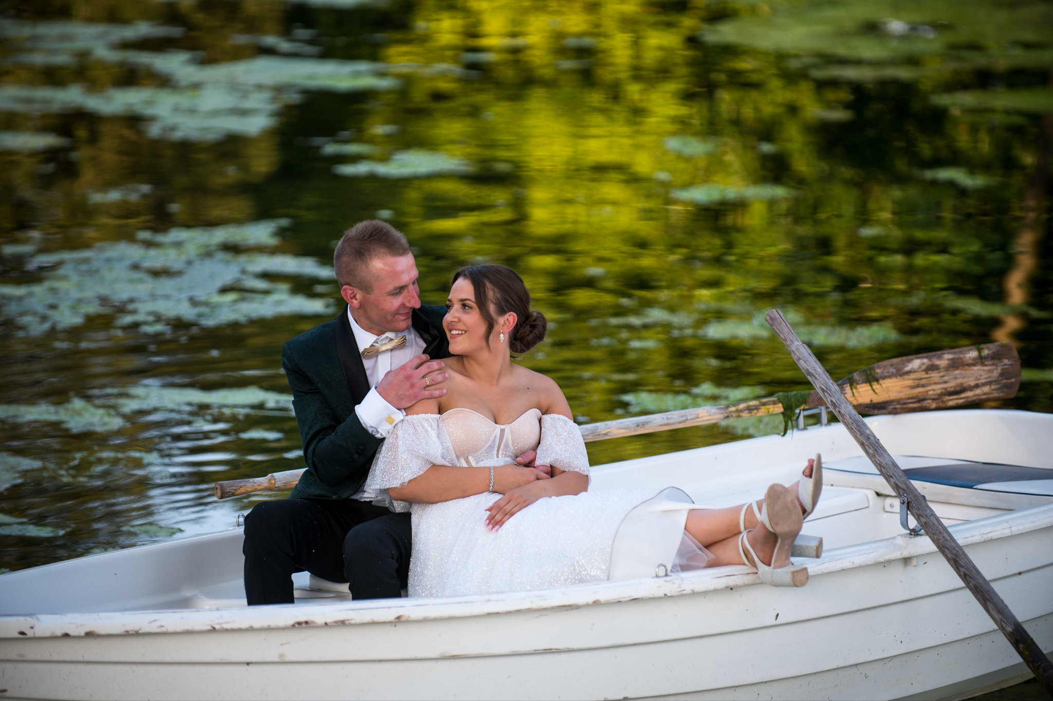 Zakochana para młoda w łódce na jeziorze, on w zielonym smokingu, ona w białej sukni, romantyczna sesja plenerowa, naturalne światło, w tle zielona woda.