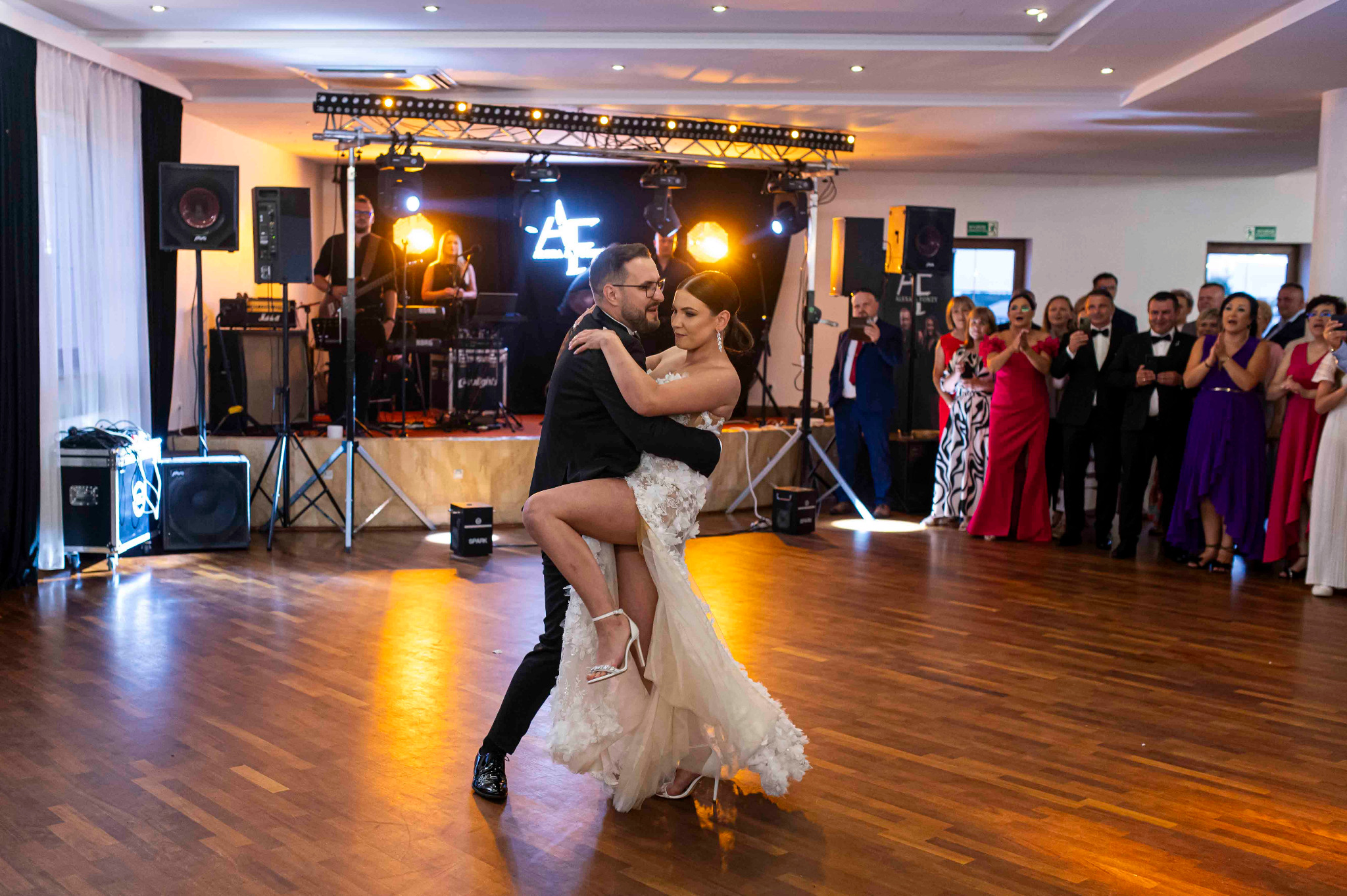Para młoda tańczy na parkiecie podczas wesela, w tle zespół muzyczny i goście. Dekoracja sali z oświetleniem i neonem. Suknia panny młodej z koronkowymi aplikacjami.